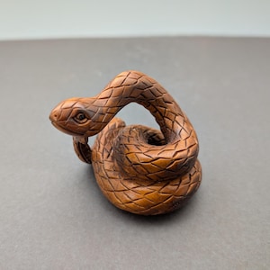 Puede incluir: Tallado en madera detallado de una serpiente en posición enrollada. La serpiente es marrón con escamas texturizadas y una cabeza detallada. El tallado se encuentra sobre un fondo gris.