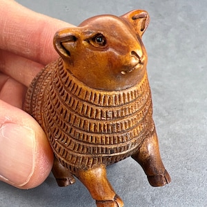 May include: A wooden carving of a sheep with a detailed, textured wool pattern. The sheep has a brown color and a realistic face with a small black eye.
