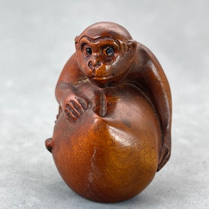 Peut inclure: Une sculpture en bois sculptée à la main d'un singe tenant un fruit rond. Le singe est brun et a les yeux noirs. Le fruit est également brun et a une surface lisse.