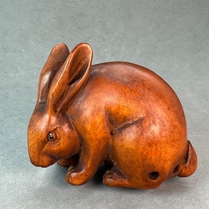 May include: A hand-carved wooden rabbit sculpture with a brown finish. The rabbit is depicted in a crouching position with its ears perked up.