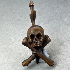 Peut inclure: Un crâne en bois avec des os croisés, le crâne est sculpté avec des détails des traits du visage. Le crâne est monté sur une base en bois.