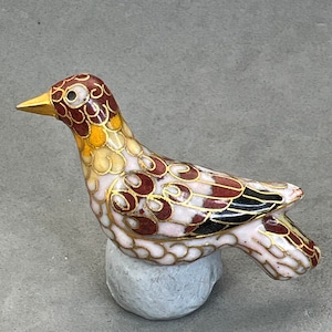 May include: A small, colourful bird figurine made of cloisonné enamel. The bird is perched on a round, white base. The bird's body is decorated with intricate patterns in shades of red, yellow, and white.