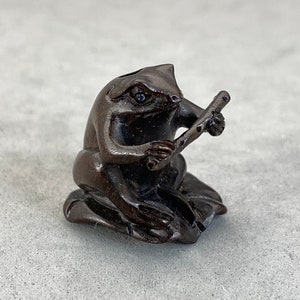 May include: A dark brown wooden frog figurine playing a flute. The frog is sitting on a leaf and has a detailed carving.