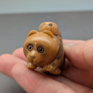 May include: A small, carved figurine of a bear with a smaller bear cub riding on its back. The figurine is a light brown color with dark brown accents and detailed facial features. The bear's eyes are dark and round, and the cub's eyes are also visible. The figurine is held in a hand.