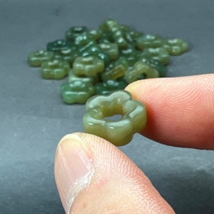 May include: A close-up of a jade green flower-shaped bead with a hole in the center. The bead is being held by a hand.
