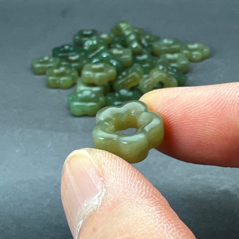 May include: A close-up of a jade green flower-shaped bead with a hole in the center. The bead is being held by a hand.