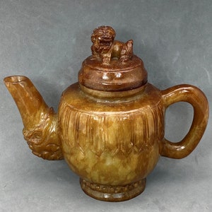 Könnte beinhalten: Eine braune und gelbe Jade-Teekanne mit einem geschnitzten Löwenkopf als Knauf. Die Teekanne hat einen runden Körper mit einem geschnitzten Design und einen Ausguss an der Seite.
