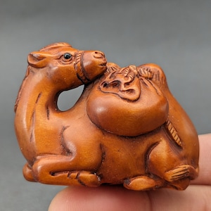 May include: A hand-carved wooden camel figurine with a large sack on its back. The camel is brown and the sack is a lighter brown. The figurine is detailed with intricate carvings.