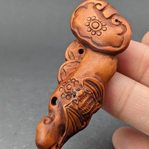 May include: A hand-carved wooden pendant featuring a bat with intricate details and a stylized flower design. The pendant is made of dark brown wood and has a smooth, polished finish.