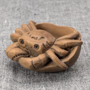 Peut inclure: Une sculpture en bois sculptée à la main d'un crabe, avec des détails précis et des yeux réalistes. Le crabe est représenté en position recroquevillée, comme s'il se cachait dans sa carapace.