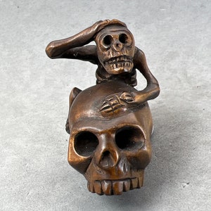 May include: A wooden sculpture of a skull with a small skeleton figure sitting on top of it. The skeleton figure has its hands on its head and is looking down at the skull.
