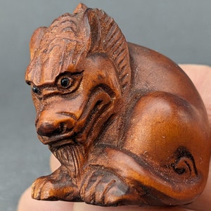 Peut inclure: Une petite sculpture en bois sculptée de manière complexe représentant une créature mythique grognante avec des yeux noirs. La sculpture est brune et présente une surface texturée détaillée.