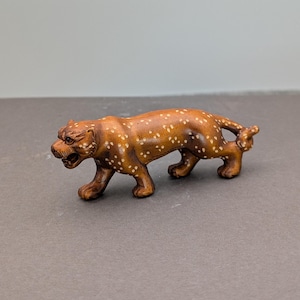 May include: A small, intricately carved wooden tiger figurine. The tiger is brown with white spots and is depicted in a walking pose. The figurine has detailed facial features and a textured surface, showcasing the craftsmanship of the carving.