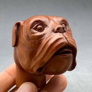 Puede incluir: Una figurita de cabeza de bulldog de madera tallada a mano. La figurita está hecha de madera marrón oscura y tiene un aspecto realista. Los ojos del bulldog están muy abiertos y su boca está ligeramente abierta.