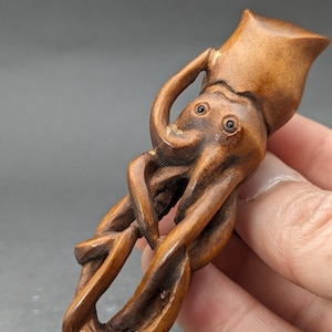 May include: A hand-carved wooden sculpture of an octopus with detailed features, including eyes and tentacles. The octopus is depicted in a curled position, with its tentacles intertwined.