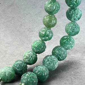 Puede incluir: Una hebra de cuentas de jade verde, cada una tallada con intrincados diseños florales. Las cuentas están enhebradas en un cordón y están dispuestas en un patrón curvo.