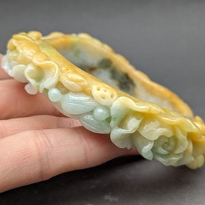 Op de afbeelding: Een jade armband gegraveerd met ingewikkelde bloemmotieven. De armband is lichtgroen met gele accenten.