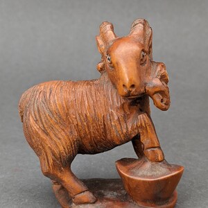 May include: A hand-carved wooden figurine of a goat standing on a small base. The goat is brown and has a detailed face with large eyes. It is holding a small, round object in its mouth.