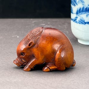 Peut inclure: Petite sculpture en bois finement sculptée représentant un sanglier. Le sanglier est représenté en position assise, la tête baissée et le groin touchant le sol. Le bois a une teinte brun-rougeâtre chaude.