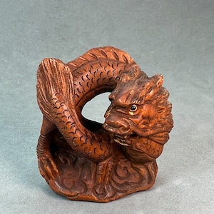 Può includere: Una scultura in legno intagliata a mano di un drago arrotolato in un cerchio. Il drago è marrone e presenta squame dettagliate e un'espressione feroce.