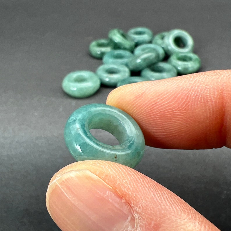May include: A close-up of a jade green donut-shaped bead with a smooth, polished surface. The bead is being held by a finger, and there are other similar beads in the background.