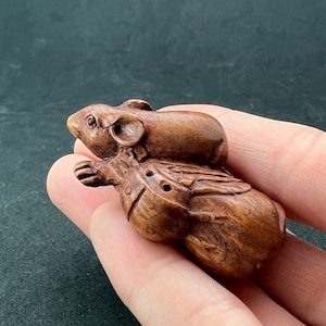 Puede incluir: Una escultura de madera tallada a mano de dos ratones. El ratón más grande está sentado de espaldas al espectador, mientras que el ratón más pequeño está encaramado en su espalda. La escultura está hecha de madera marrón oscura y tiene un acabado natural.