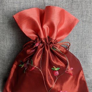 May include: A red drawstring bag with a coral colored top and floral embroidery. The bag is made of satin and has a gathered top.
