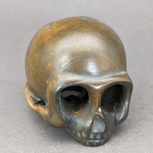 May include: A hand-carved wooden skull sculpture with a dark brown finish. The skull has a detailed face with eye sockets, a nose, and teeth.