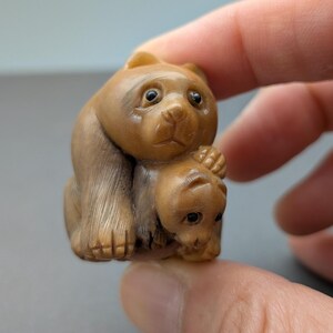 May include: A carved wooden figurine of a bear and cub. The bear is sitting with the cub nestled in its arms. The figurine is a light brown color and has detailed features, including the bear's fur and the cub's face.