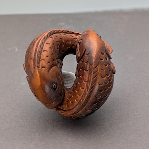 May include: A carved wooden sculpture depicting two fish intertwined in a circular form. The fish are detailed with scales and fins, and have dark eyes. The wood has a warm, brown tone, and the sculpture is set against a neutral background.