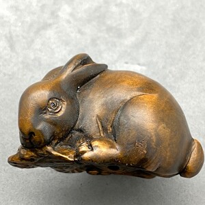 Peut inclure: Une figurine de lapin en bois sculpté à la main avec une finition brune et dorée. Le lapin est représenté en position assise, la tête inclinée sur le côté. La figurine est détaillée avec des traits réalistes, notamment un long nez, de petites oreilles et une queue touffue.