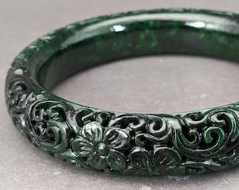 May include: A dark green jade bangle with intricate floral carvings. The bangle is polished and has a smooth surface.