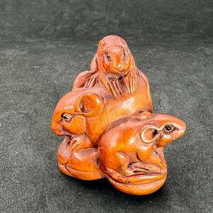 Puede incluir: Una escultura de madera tallada a mano de tres ratones. Los ratones son marrones y tienen rasgos detallados. La escultura es un netsuke japonés tradicional.