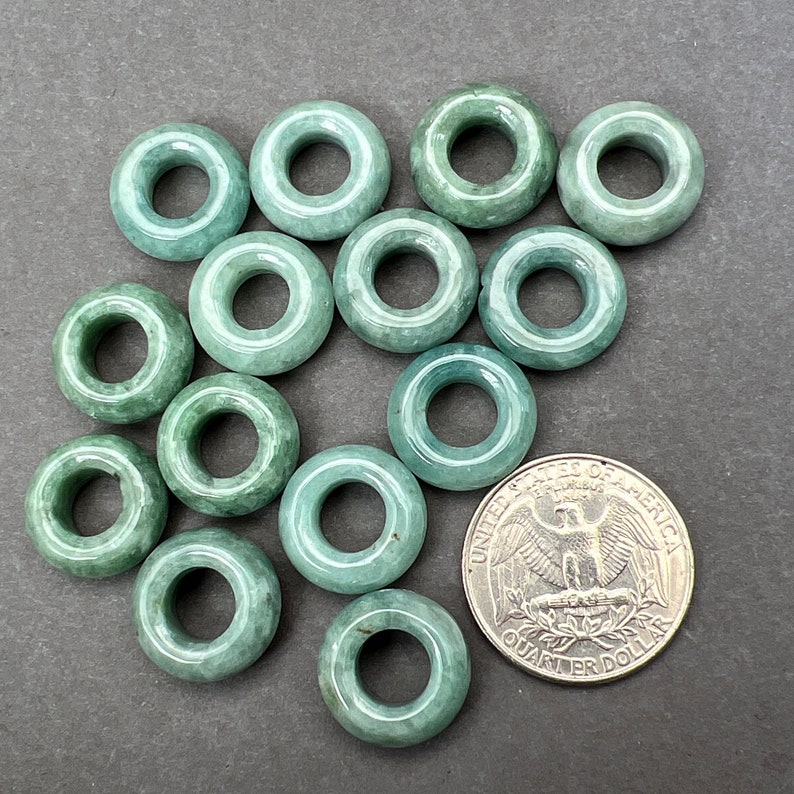 May include: A collection of 14 jade green donut-shaped beads with a hole in the center. The beads are arranged in a random pattern on a gray surface. A US quarter is included for size reference.