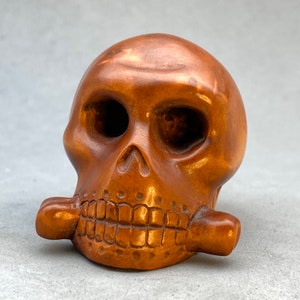 May include: A wooden sculpture of a skull with a carved wooden pipe in its mouth. The skull is brown and has a detailed carving of teeth.