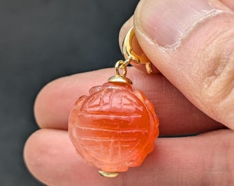 May include: A gold pendant with a carved carnelian stone in the shape of a sphere. The stone is a deep orange color and has intricate carvings on its surface.