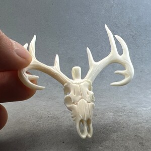 May include: Ivory-colored deer skull pendant with detailed antlers. The skull is carved with visible eye sockets and nasal cavity. The antlers extend outward, showcasing the intricate design of the pendant. The pendant is held against a gray background.