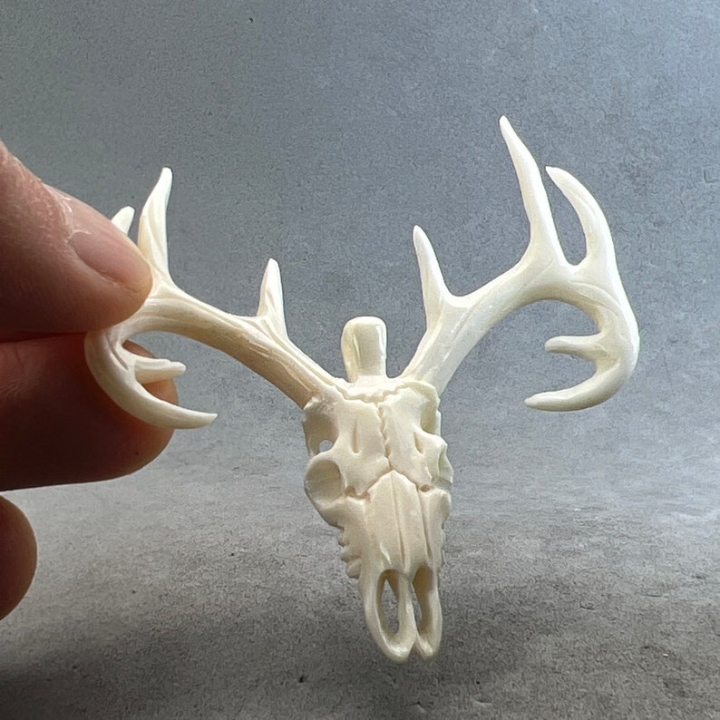 May include: Ivory-colored deer skull pendant with detailed antlers. The skull is carved with visible eye sockets and nasal cavity. The antlers extend outward, showcasing the intricate design of the pendant. The pendant is held against a gray background.