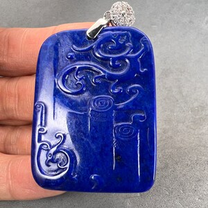 Op de afbeelding: Een diepblauwe lapis lazuli hanger met ingewikkelde gravures van een draak en rollen. De hanger is gezet in zilver en heeft een klein, helder kristal aan de bovenkant.