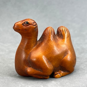 May include: A hand-carved wooden camel figurine with a brown finish. The camel is sitting with its legs tucked underneath its body. The camel has a hump on its back and a long neck.