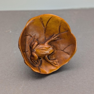 Peut inclure: Une grenouille en bois sculpté reposant sur une feuille de nénuphar. La grenouille a des pattes et des yeux détaillés. La feuille de nénuphar a des veines détaillées et une forme incurvée. Le bois a une teinte brune chaude.