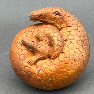 May include: A wooden carving of a pangolin curled up with its young. The carving is detailed and shows the scales of the pangolin.