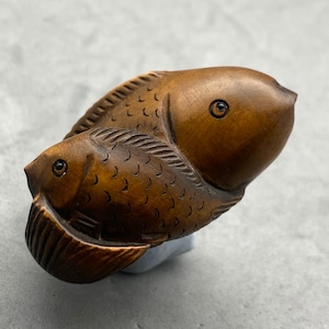 Peut inclure: Deux poissons en bois sculptés de manière complexe, l'un plus petit que l'autre, sont assemblés. Le plus grand poisson est brun avec un œil brun foncé, tandis que le plus petit poisson est brun clair avec un œil brun foncé. Les deux poissons ont des écailles et des nageoires détaillées.