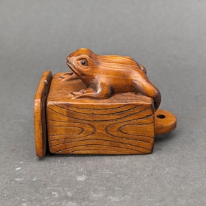 May include: A detailed wooden carving of a frog perched on a rectangular wooden box. The frog is brown with visible eyes and sits atop a box with wood grain details. The box has a small, circular handle on the side.