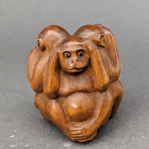 May include: A wooden sculpture of three monkeys in a seated position. Each monkey is in a different pose, covering its eyes, ears, and mouth. The sculpture is carved from a light brown wood and has a smooth finish.