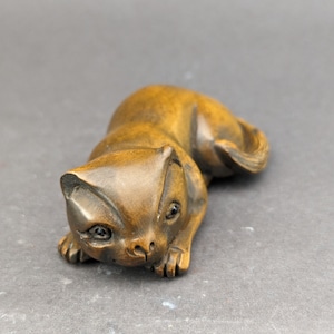 May include: A small, intricately carved wooden sculpture of a cat lying down. The cat is brown and has a detailed face with black eyes.