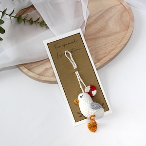 Könnte beinhalten: Ein kleines, weißes, gehäkeltes Möwenornament mit einem grauen Flügel, einem orangefarbenen Schnabel und orangefarbenen Füßen. Die Möwe ist an einer weißen Schnur befestigt und hat einen kleinen rot-weißen Rettungsring. Sie ist auf einer rechteckigen Karte montiert.