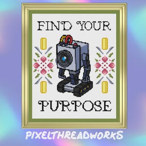 May include: Cross-stitch art in a gold frame. The design features a robot with the text "FIND YOUR PURPOSE". The robot is gray with black details and a large lens. Floral and geometric patterns surround the robot.