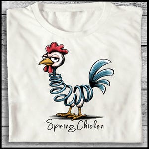 May include: White t-shirt featuring a cartoon rooster with a spring-like body in blue. The rooster has a red comb and wattle, a yellow beak, and a blue tail. The text "Spring Chicken" is written below the rooster in a script font.