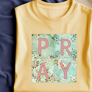 Könnte beinhalten: Gelbes T-Shirt mit floralem Muster und dem Wort "PRAY" in rosa Buchstaben.